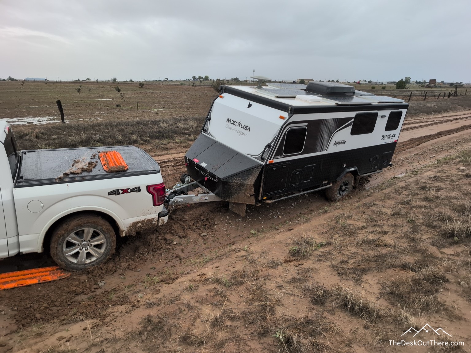 White pickup and off-road camper stuck in a muddy rut with orange recovery boards.