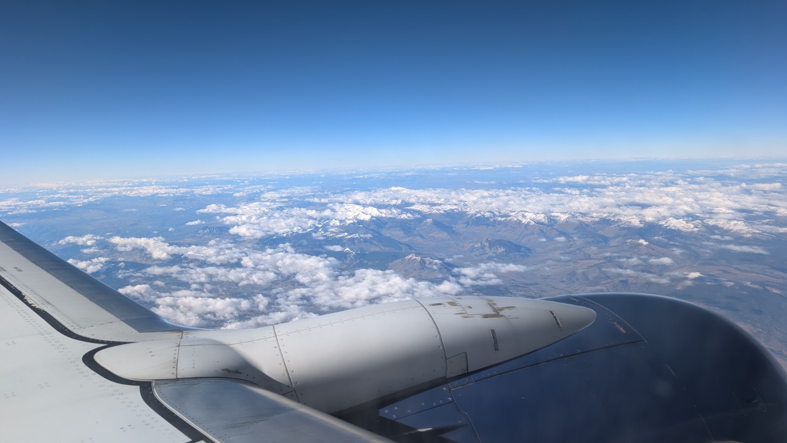 View from an airplane window of snow-capped mountains above scattered clouds, with the wing and engine in the foreground.