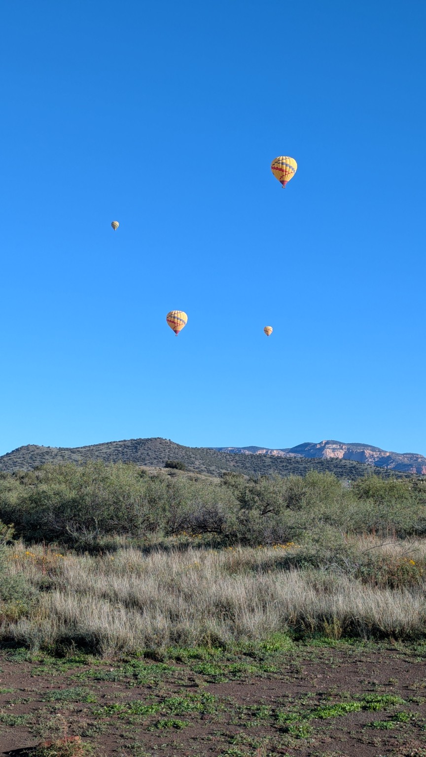 Four colorful hot air balloons floating above scrub-covered Arizona hills under a clear blue sky.